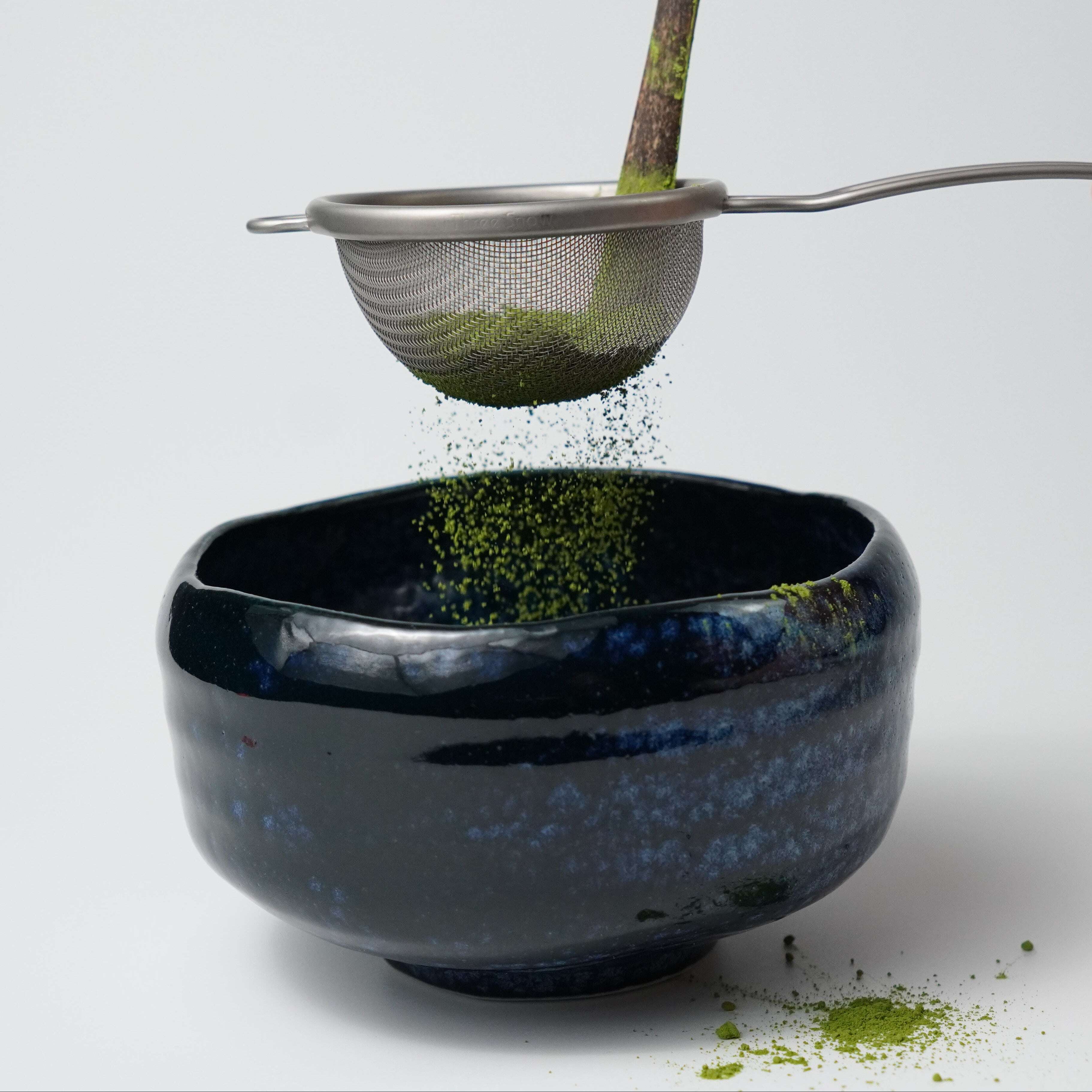 Sifting green Koharu no Shiro superior matcha powder from Yamada Chaya into a dark blue ceramic tea bowl with a made in Japan steel sieve.