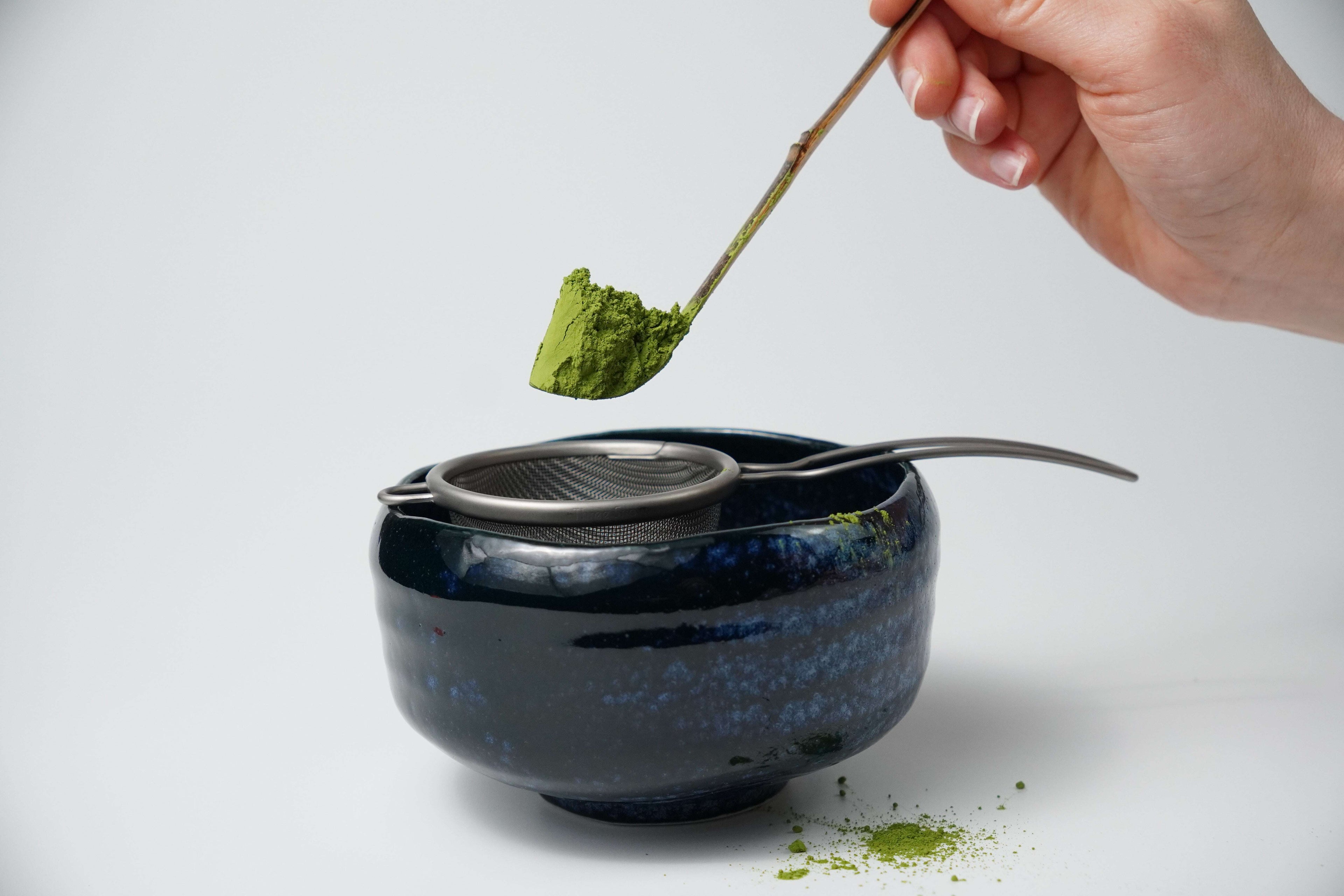 Person holding a matcha scoop with green Koharu no Shiro matcha powder by Yamada Chaya over a blue ceramic bowl on a white background